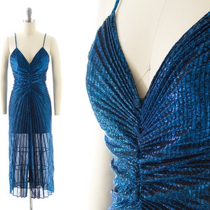Op de afbeelding: Een glinsterende blauwe jurk met spaghettibandjes en een V-hals. De jurk heeft een geplooid lijfje en een doorschijnende, vloerlange rok. De stof heeft een getextureerde, metallic uitstraling, wat een glamoureuze look creëert.