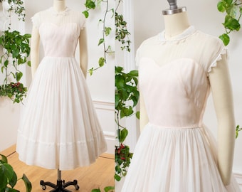 May include: A vintage-style, knee-length dress in soft pink and white. The bodice is a solid pink, with a sweetheart neckline and sheer overlay. The skirt is a flowing white, with a delicate lace trim at the hem. The dress is displayed on a mannequin.