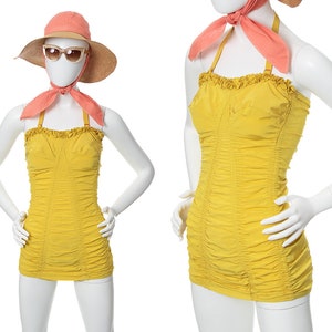 May include: A yellow swimsuit with a ruffled neckline and a gathered skirt. The swimsuit is on a white mannequin with a pink scarf and a straw hat.
