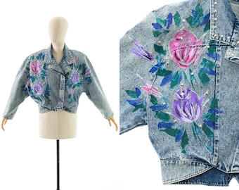 May include: A light blue denim jacket with a cropped fit and floral embroidery. The flowers are pink, purple, and blue with green leaves. The jacket has a zip closure and a stand-up collar.