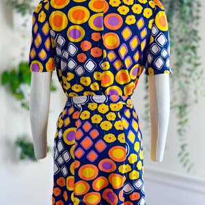 May include: A vintage dress with a striking geometric pattern. The navy blue dress is decorated with orange, yellow, purple, and white shapes. The short-sleeved dress is displayed on a mannequin, highlighting its retro design.
