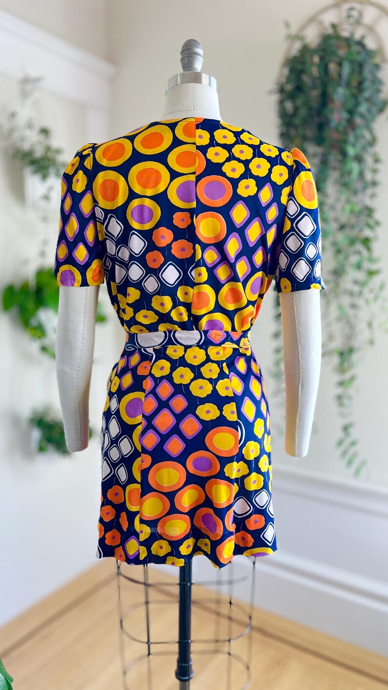 May include: A vintage dress with a striking geometric pattern. The navy blue dress is decorated with orange, yellow, purple, and white shapes. The short-sleeved dress is displayed on a mannequin, highlighting its retro design.