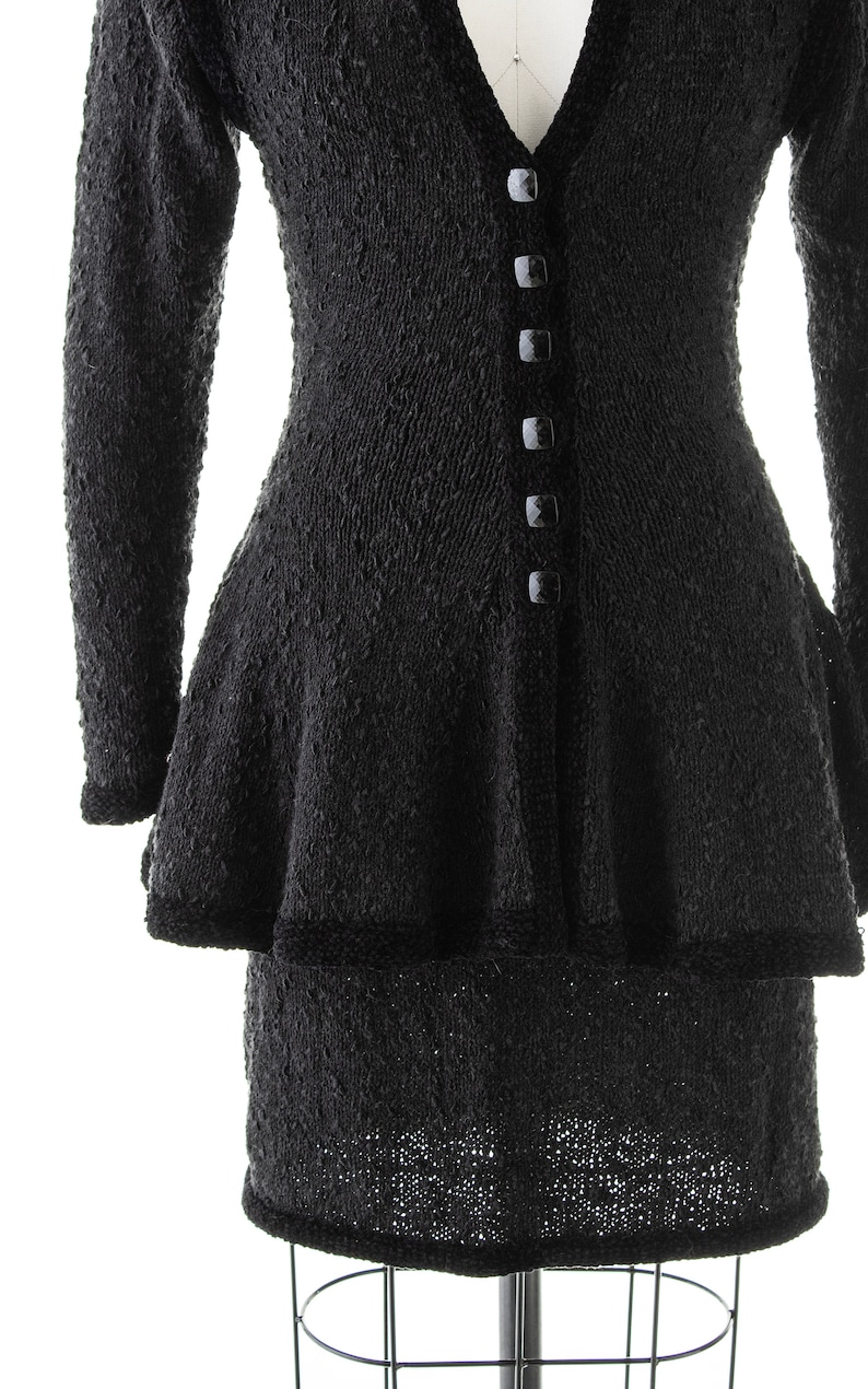 May include: Black knitted dress with a fitted bodice and a flared skirt. The dress has a V-neckline and a row of black buttons down the front.