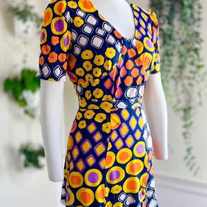 May include: A retro-style dress with a bold geometric design. The dress has a V-neck, short sleeves, and a cinched waist. The pattern incorporates orange, yellow, purple, and white shapes set against a navy blue backdrop. The dress is displayed on a mannequin.