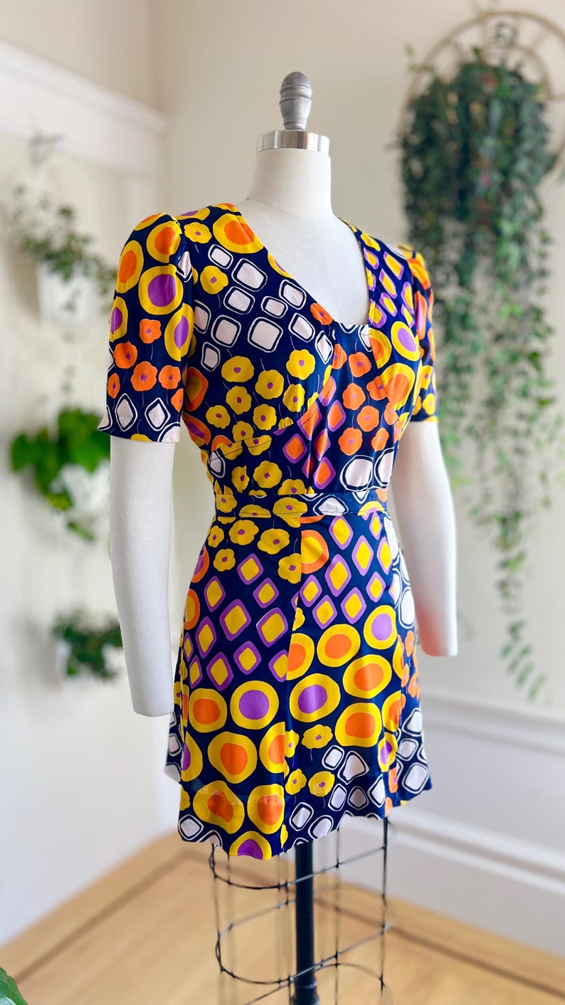May include: A retro-style dress with a bold geometric design. The dress has a V-neck, short sleeves, and a cinched waist. The pattern incorporates orange, yellow, purple, and white shapes set against a navy blue backdrop. The dress is displayed on a mannequin.