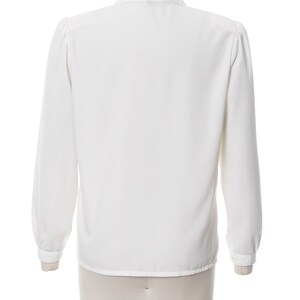Puede incluir: Una blusa blanca de manga larga con cuello fruncido y manga ligeramente abullonada. La blusa est&aacute; en un maniqu&iacute; blanco.
