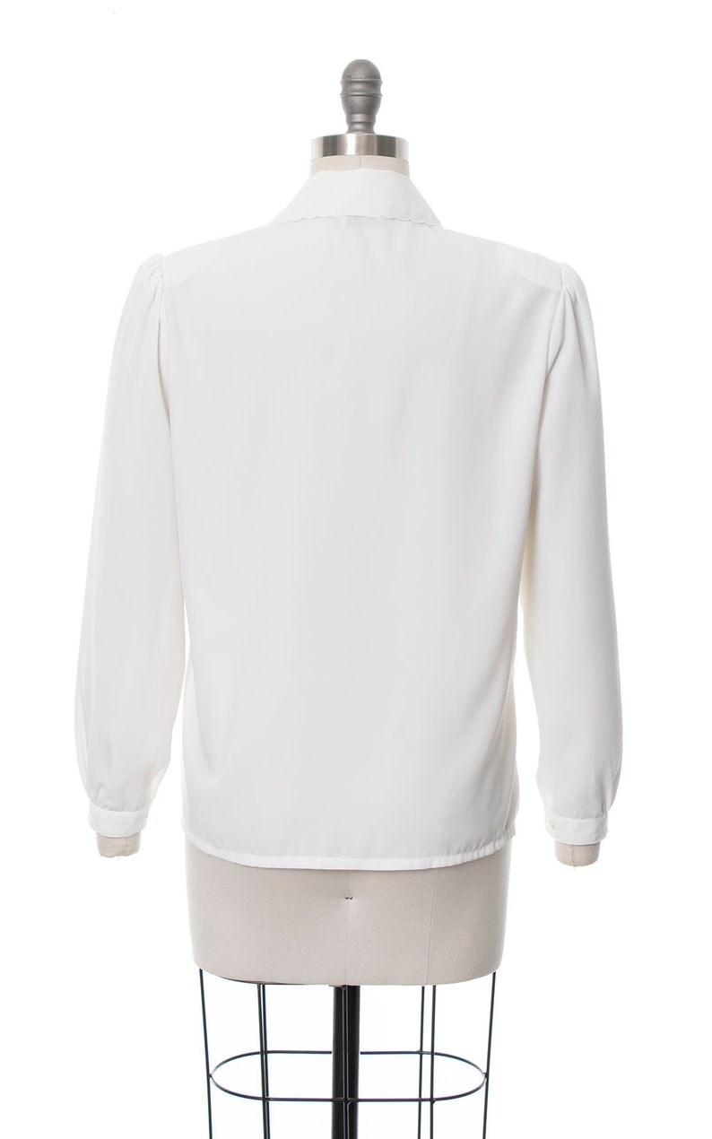Puede incluir: Una blusa blanca de manga larga con cuello fruncido y manga ligeramente abullonada. La blusa est&aacute; en un maniqu&iacute; blanco.