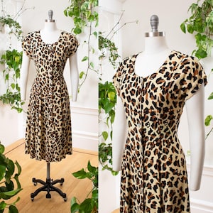 May include: A vintage leopard print dress with a scoop neckline and short sleeves. The dress features a button-down front and a fitted waist, with a flared skirt. The dress is displayed on a mannequin against a backdrop of greenery.