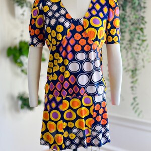 May include: A vintage dress with a V-neck and short sleeves, displayed on a mannequin. The dress features a vibrant pattern of orange, yellow, purple, and white geometric shapes on a navy blue background. The dress has a flared skirt.