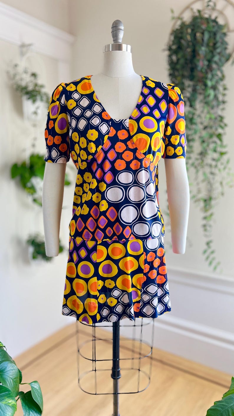 May include: A vintage dress with a V-neck and short sleeves, displayed on a mannequin. The dress features a vibrant pattern of orange, yellow, purple, and white geometric shapes on a navy blue background. The dress has a flared skirt.
