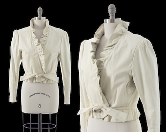 May include: A white wrap jacket with a ruffled collar and a tie at the waist. The jacket has long sleeves and a cropped length. The jacket is on a white mannequin.