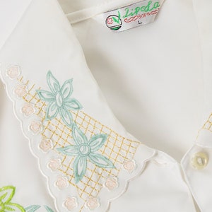 Puede incluir: Camisa blanca bordada con cuello festoneado y dise&ntilde;o floral. La camisa tiene un dise&ntilde;o de hojas y flores verdes con un patr&oacute;n de cuadr&iacute;cula dorada. La etiqueta de la camisa dice "Lipela".