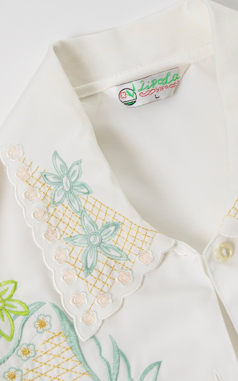 Puede incluir: Camisa blanca bordada con cuello festoneado y dise&ntilde;o floral. La camisa tiene un dise&ntilde;o de hojas y flores verdes con un patr&oacute;n de cuadr&iacute;cula dorada. La etiqueta de la camisa dice "Lipela".