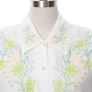 Puede incluir: Una camisa blanca de botones con cuello festoneado y dise&ntilde;o floral bordado. Las flores son verdes y azules con un contorno amarillo.