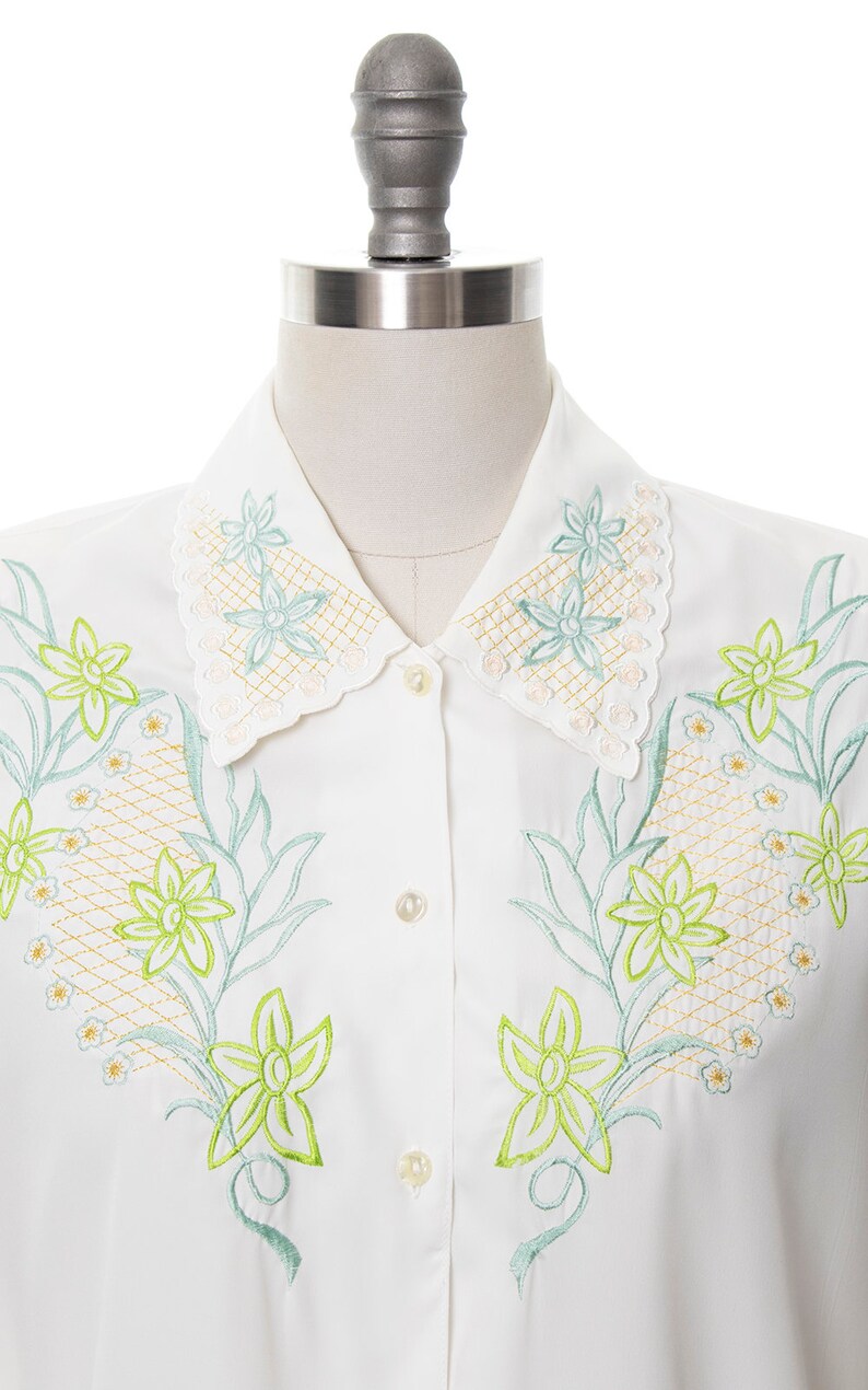 Puede incluir: Una camisa blanca de botones con cuello festoneado y dise&ntilde;o floral bordado. Las flores son verdes y azules con un contorno amarillo.