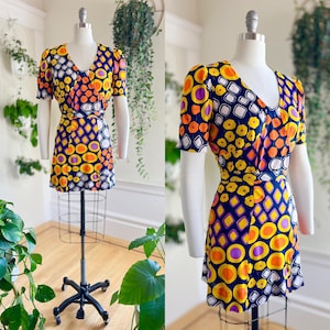 May include: A vintage mini dress with a V-neck and short sleeves. The dress has a vibrant pattern of orange, yellow, purple, and white geometric shapes on a navy blue background. It has a matching belt and is displayed on a mannequin.