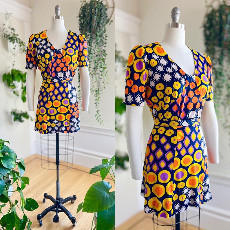May include: A vintage mini dress with a V-neck and short sleeves. The dress has a vibrant pattern of orange, yellow, purple, and white geometric shapes on a navy blue background. It has a matching belt and is displayed on a mannequin.