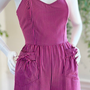 May include: A pink and black striped jumpsuit with thin straps and two pockets on the front. The pockets have a bow detail.