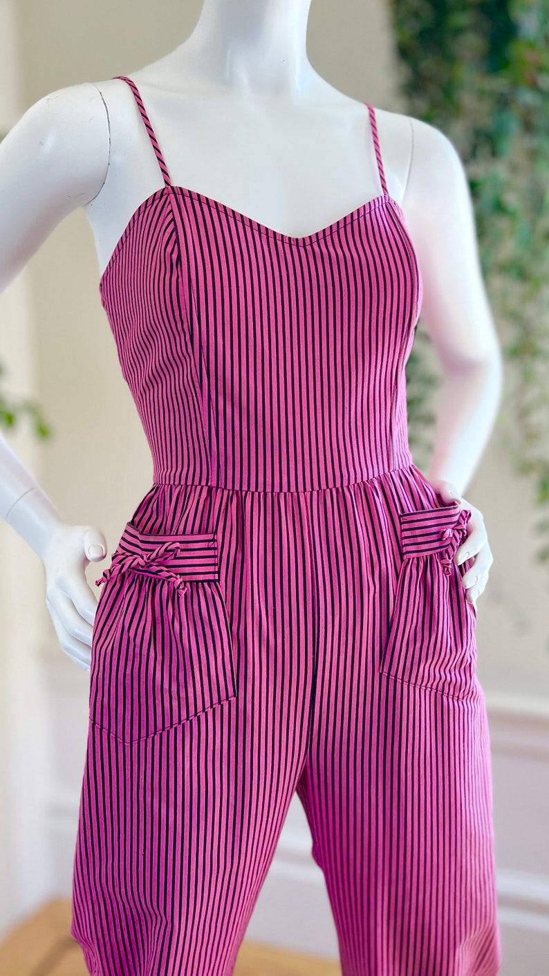 May include: A pink and black striped jumpsuit with thin straps and two pockets on the front. The pockets have a bow detail.