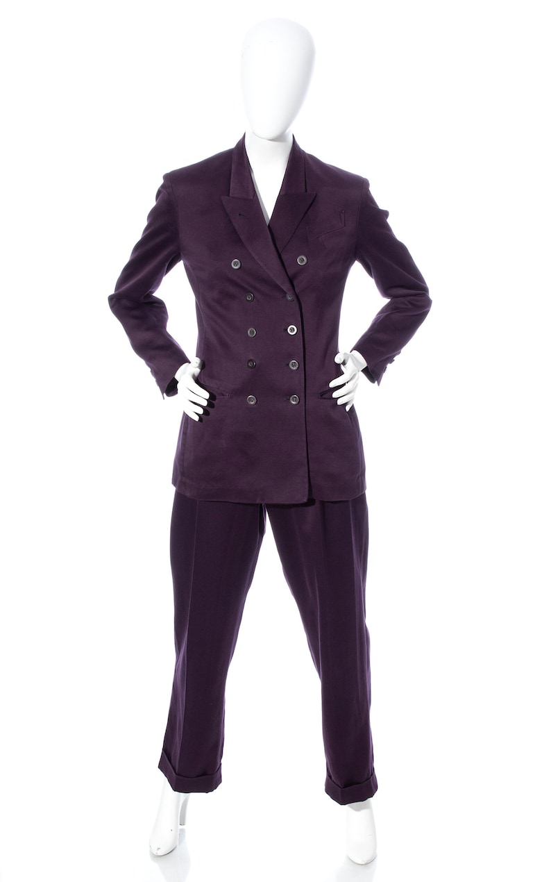 Pu&ograve; includere: Un abito doppiopetto di colore viola scuro. Il completo comprende una giacca con revers a punta e un paio di pantaloni abbinati.