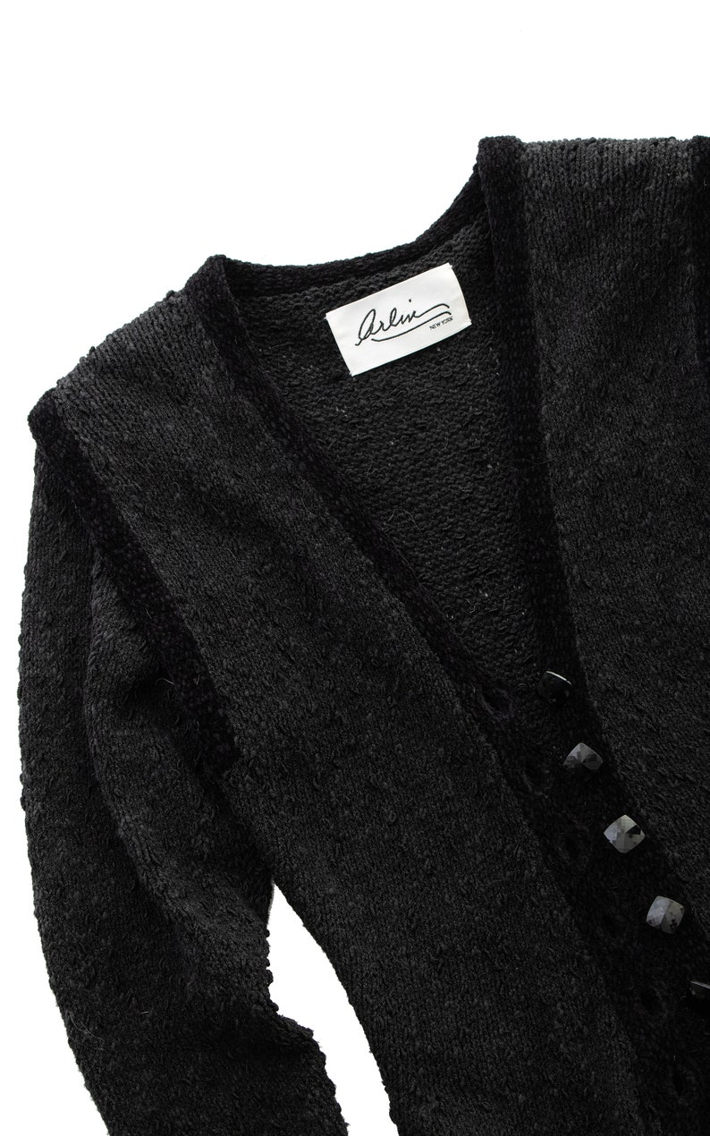 May include: Black long-sleeved cardigan with a V-neck and button closure. The cardigan has a textured knit and a label that reads "Arlini New York".