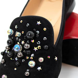 May include: Black suede loafers with a star and jewel embellishment. The loafers have a pointed toe and a low heel.