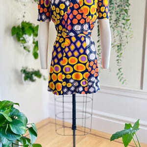 May include: A vintage mini dress with a vibrant geometric pattern in yellow, orange, purple, and white on a navy blue background. The dress features a V-neck, short sleeves, and a fitted waist. The dress is displayed on a mannequin.