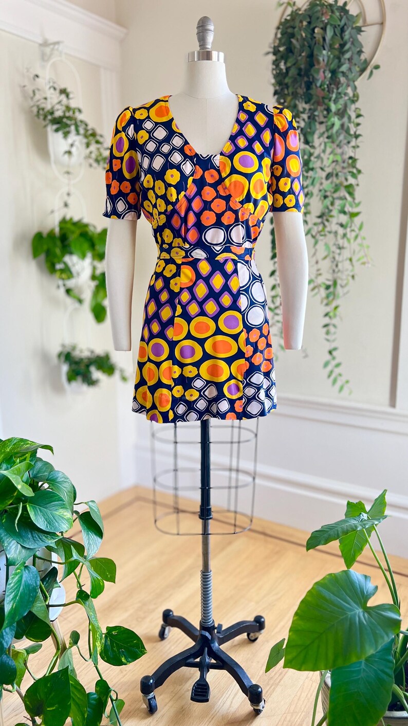 May include: A vintage mini dress with a vibrant geometric pattern in yellow, orange, purple, and white on a navy blue background. The dress features a V-neck, short sleeves, and a fitted waist. The dress is displayed on a mannequin.