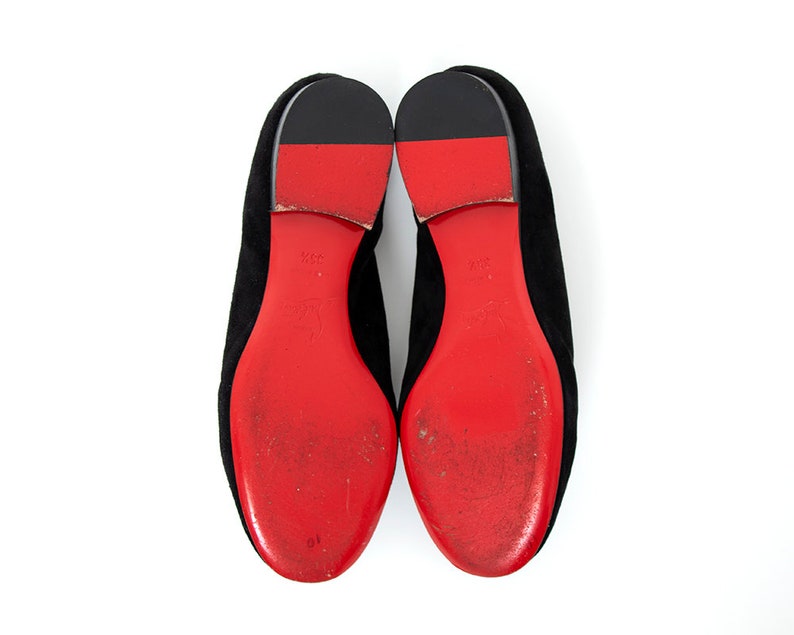 May include: A pair of black suede flats with red leather soles. The soles have a distinctive red color and a slightly worn appearance.
