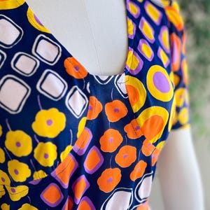 May include: A patterned dress with a V-neck design. The fabric has a repeating pattern of yellow flowers, orange circles, white squares, and purple accents on a navy blue background. The dress is displayed on a mannequin.