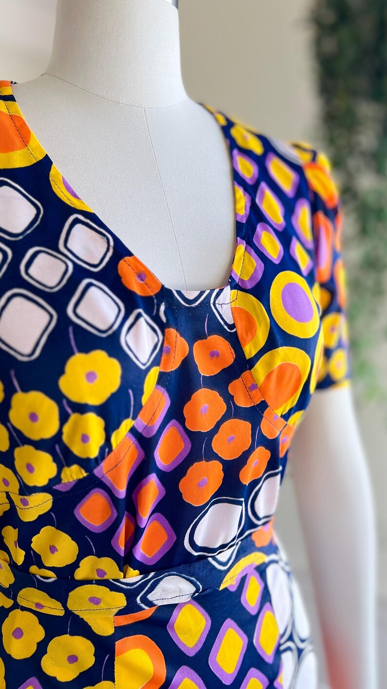 May include: A patterned dress with a V-neck design. The fabric has a repeating pattern of yellow flowers, orange circles, white squares, and purple accents on a navy blue background. The dress is displayed on a mannequin.
