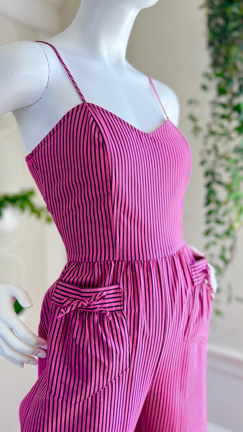 May include: Pink and black striped jumpsuit with a sweetheart neckline and two pockets. The pockets have a decorative bow detail.