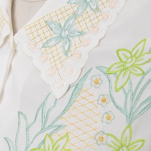 Puede incluir: Camisa blanca con un delicado dise&ntilde;o de bordado floral. El bordado presenta flores verdes y azul claro con detalles dorados. El cuello tiene un borde festoneado con un dise&ntilde;o floral m&aacute;s peque&ntilde;o.