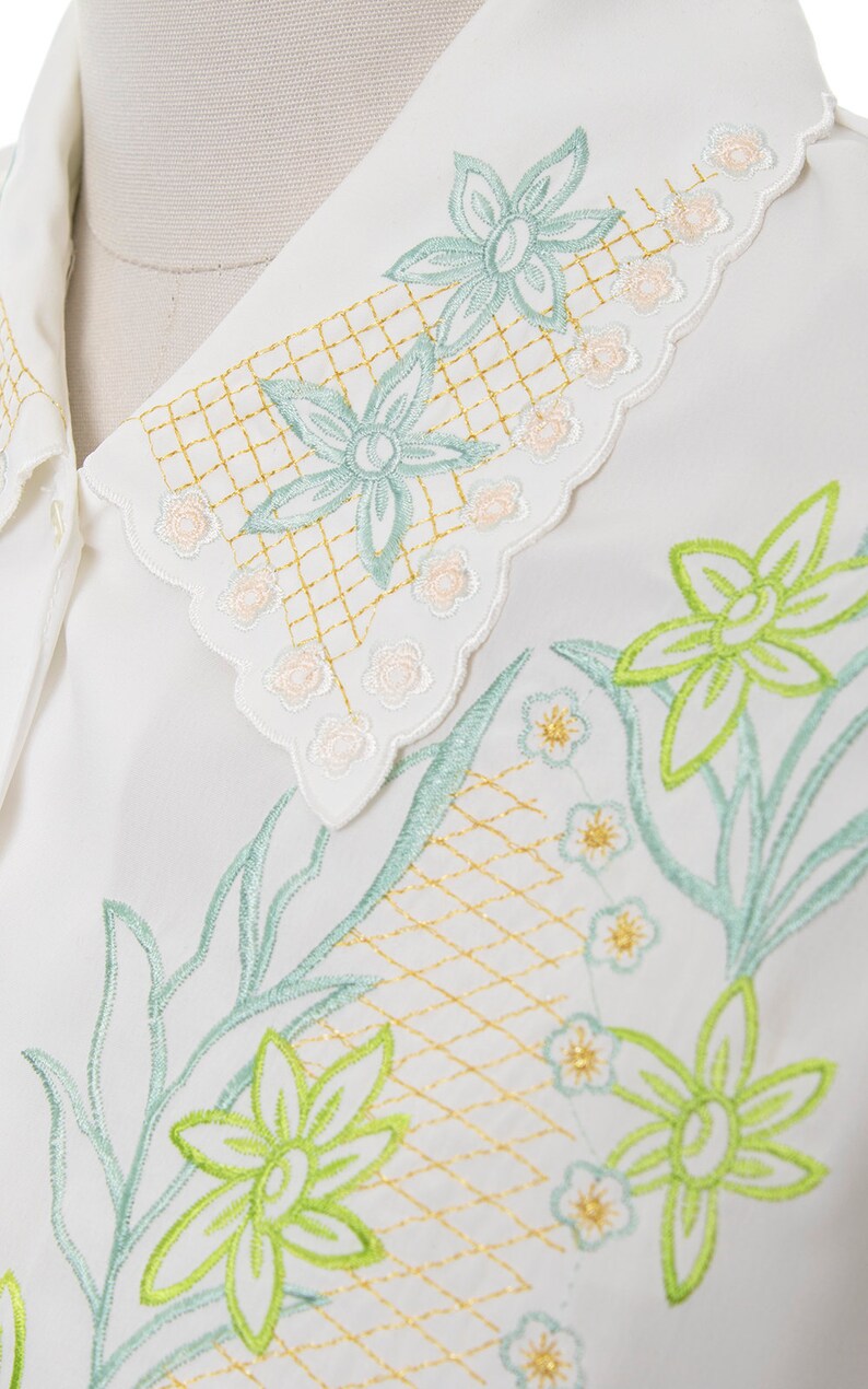 Puede incluir: Camisa blanca con un delicado dise&ntilde;o de bordado floral. El bordado presenta flores verdes y azul claro con detalles dorados. El cuello tiene un borde festoneado con un dise&ntilde;o floral m&aacute;s peque&ntilde;o.