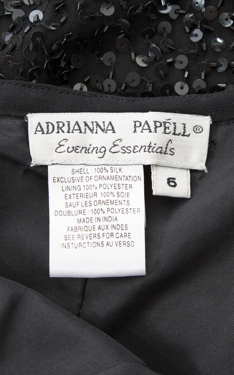 Puede incluir: Una etiqueta blanca con texto negro que dice "ADRIANNA PAPELL Evening Essentials" y enumera el contenido de la tela y las instrucciones de cuidado. La etiqueta est&aacute; unida a una prenda negra con lentejuelas.