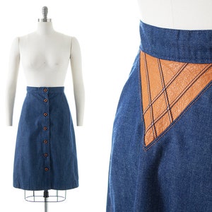 May include: A denim skirt with a brown leather panel and button closure. The skirt is a midi length and has a straight silhouette.
