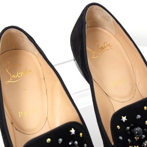 May include: A pair of black suede loafers with a star-shaped embellishment. The loafers have a gold inscription that reads "Christian Louboutin Paris".