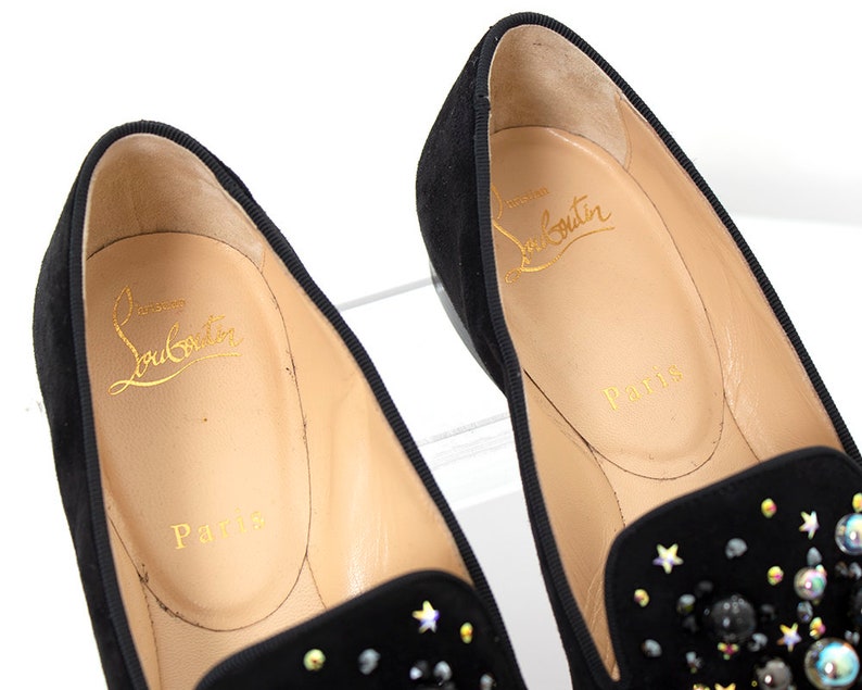 May include: A pair of black suede loafers with a star-shaped embellishment. The loafers have a gold inscription that reads "Christian Louboutin Paris".