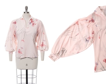 May include: A pale pink blouse with a floral print featuring a light gray background and pink, red, and blue flowers. The blouse has a V-neckline and long, gathered sleeves.