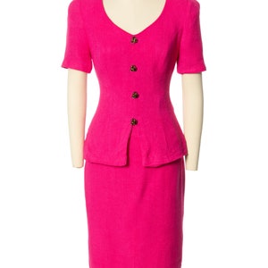 May include: A hot pink skirt suit with a fitted bodice and a skirt that falls to the knee. The suit has a V-neckline and short sleeves. The suit is fastened with gold buttons.