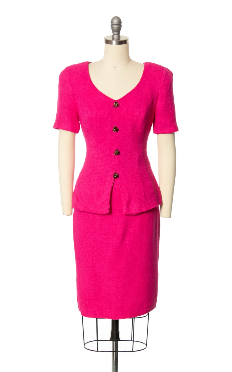 May include: A hot pink skirt suit with a fitted bodice and a skirt that falls to the knee. The suit has a V-neckline and short sleeves. The suit is fastened with gold buttons.