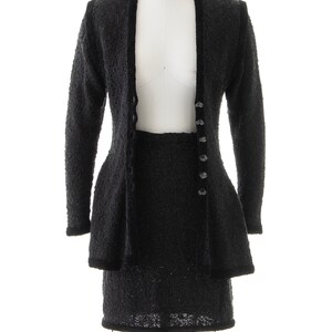 May include: A black knit skirt suit with a long-sleeved jacket and a skirt. The jacket has a V-neck and buttons down the front. The skirt is knee-length and has a fitted waist.