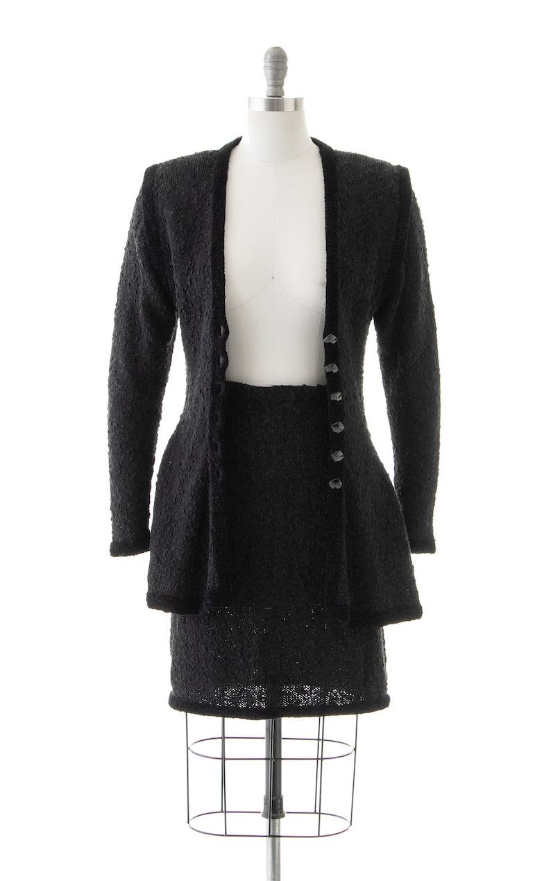 May include: A black knit skirt suit with a long-sleeved jacket and a skirt. The jacket has a V-neck and buttons down the front. The skirt is knee-length and has a fitted waist.