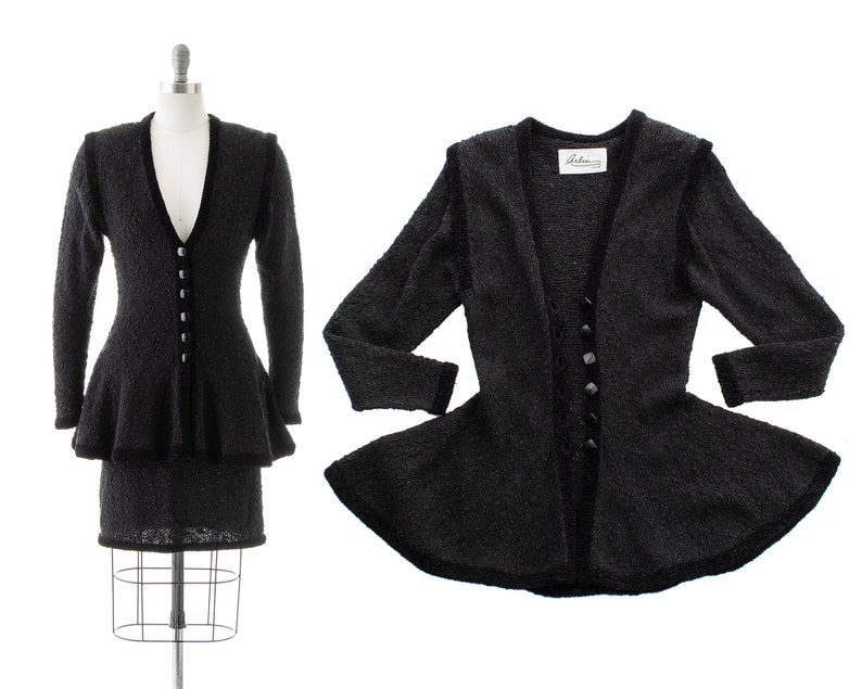 May include: A black knit suit with a fitted skirt and a peplum jacket. The jacket has a V-neckline and a button closure. The label "Adele" is visible on the jacket.