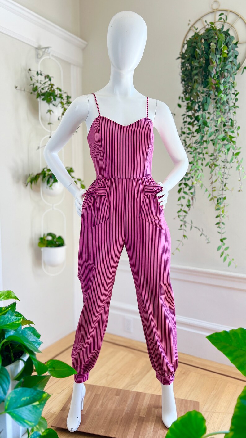 May include: A pink and black striped jumpsuit with a sweetheart neckline and cuffed legs. The jumpsuit has pockets on the front and is on a white mannequin.