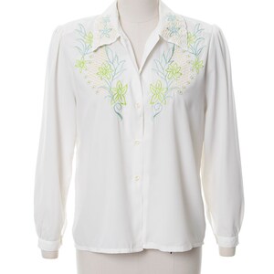 Puede incluir: Una camisa blanca de manga larga con botones y un dise&ntilde;o de bordado floral en el cuello y la parte delantera. El bordado es en tonos verdes y amarillos.