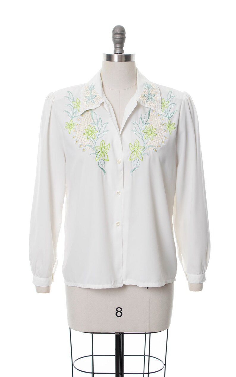 Puede incluir: Una camisa blanca de manga larga con botones y un dise&ntilde;o de bordado floral en el cuello y la parte delantera. El bordado es en tonos verdes y amarillos.