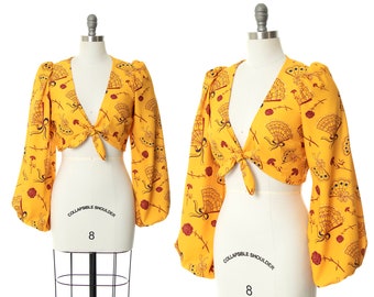 May include: A yellow crop top with a tie front and long sleeves. The top has a print of red and brown fans on a yellow background. The top is on a white mannequin.