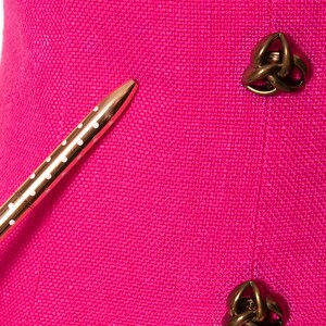 May include: A close-up of a pink fabric with two gold-toned decorative buttons. The buttons are in the shape of a knot. A gold pen with white polka dots is lying on the fabric.