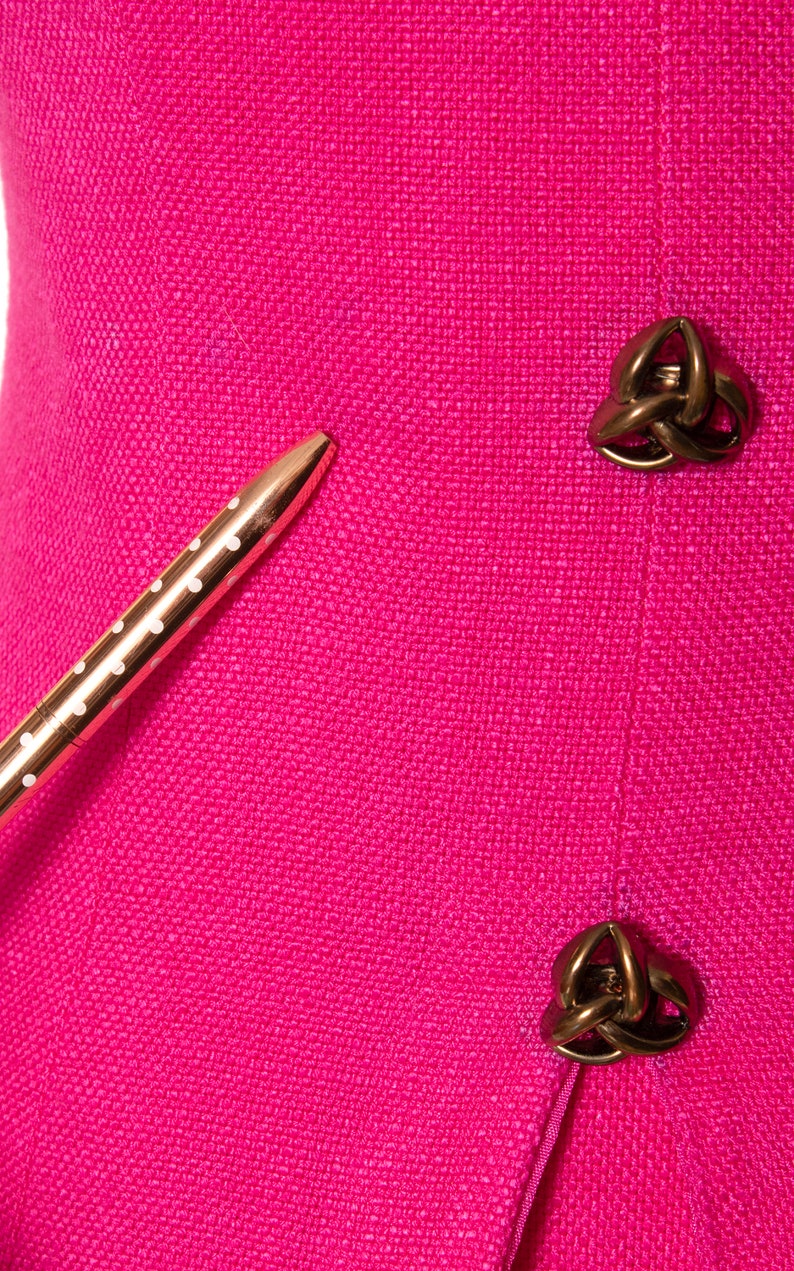 May include: A close-up of a pink fabric with two gold-toned decorative buttons. The buttons are in the shape of a knot. A gold pen with white polka dots is lying on the fabric.